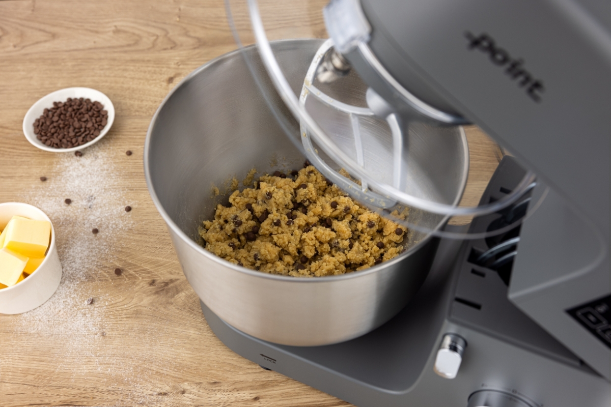 Grey-colored Point POKM5010 kitchen machine on a wooden table pictured slightly from above with cookie dough showing inside the bowl and some flour and butter on the table next to the machine