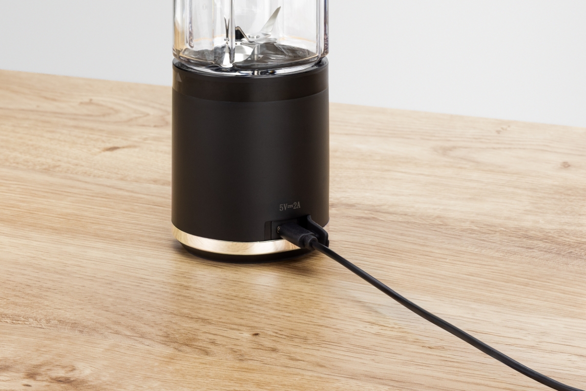 POINT POSB5000 RECHARGABLE SPORT BLENDER A black Point rechargeable travel blender on a wooden table with only its base showing and being charged with an USB-C cable