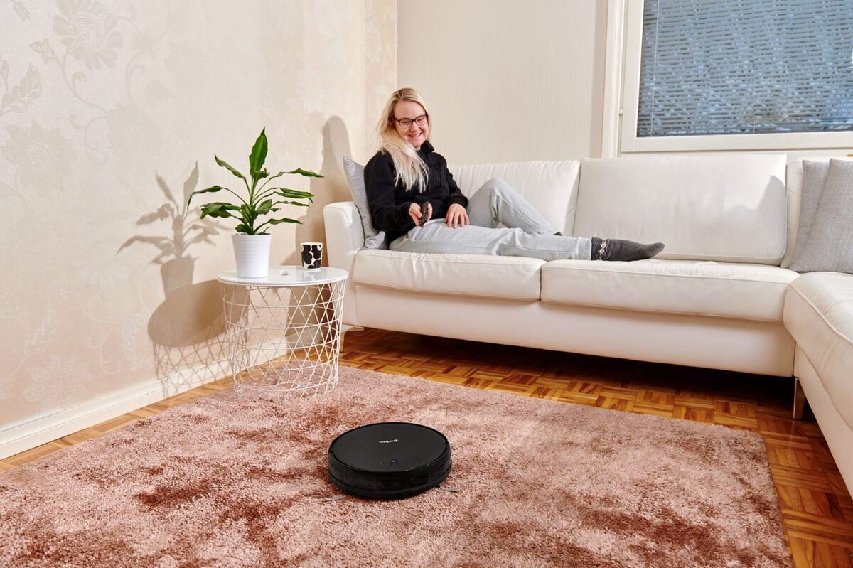 A woman sitting on a white couch in a living room and pointing the Point Dusty vacuum cleaner with a remote control and the vacuum cleaner is on a pink carpet