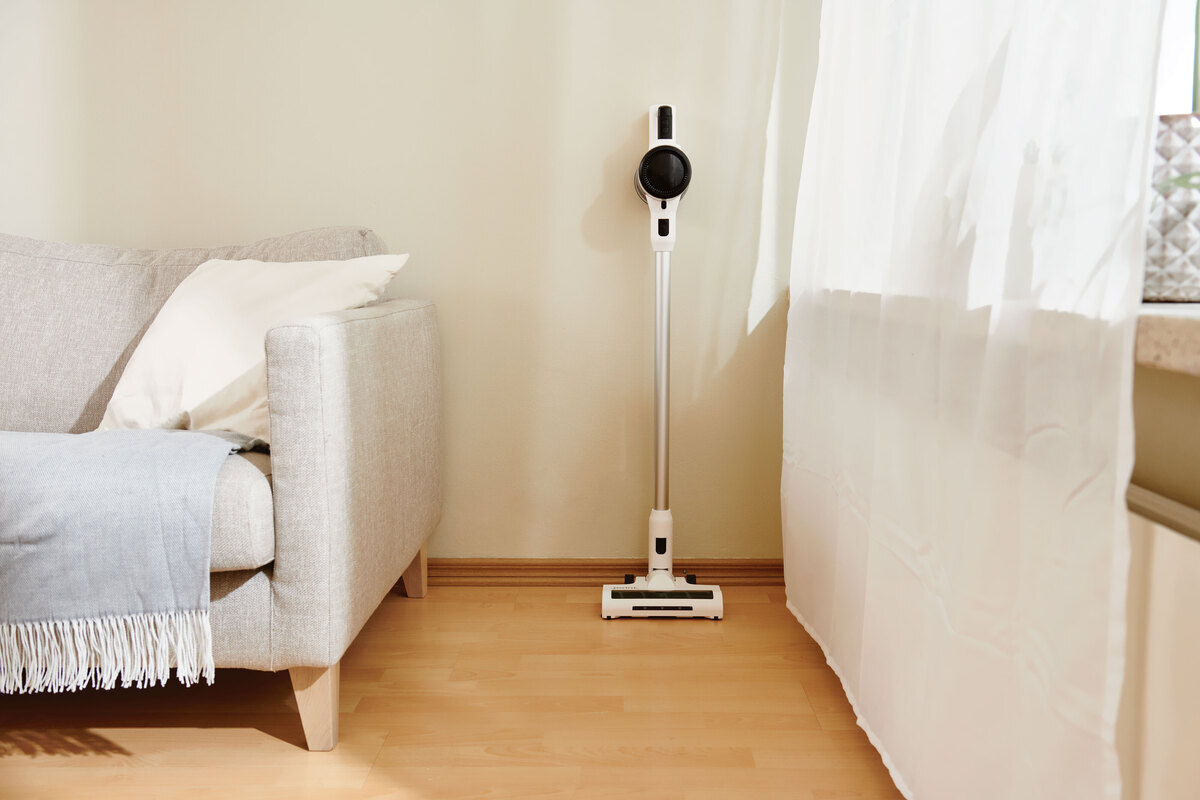 A wide angle image of the Point Pencil stick vacuum in front of a beige wall with a white sofa next to it