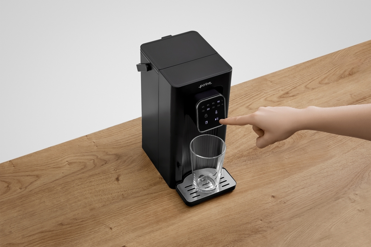 POINT POWD5000 INSTANT HOT WATER DISPENS The black Point hot water dispenser on a wooden table with a see-through glass placed on the mug holder and a hand pressing a function on the screen