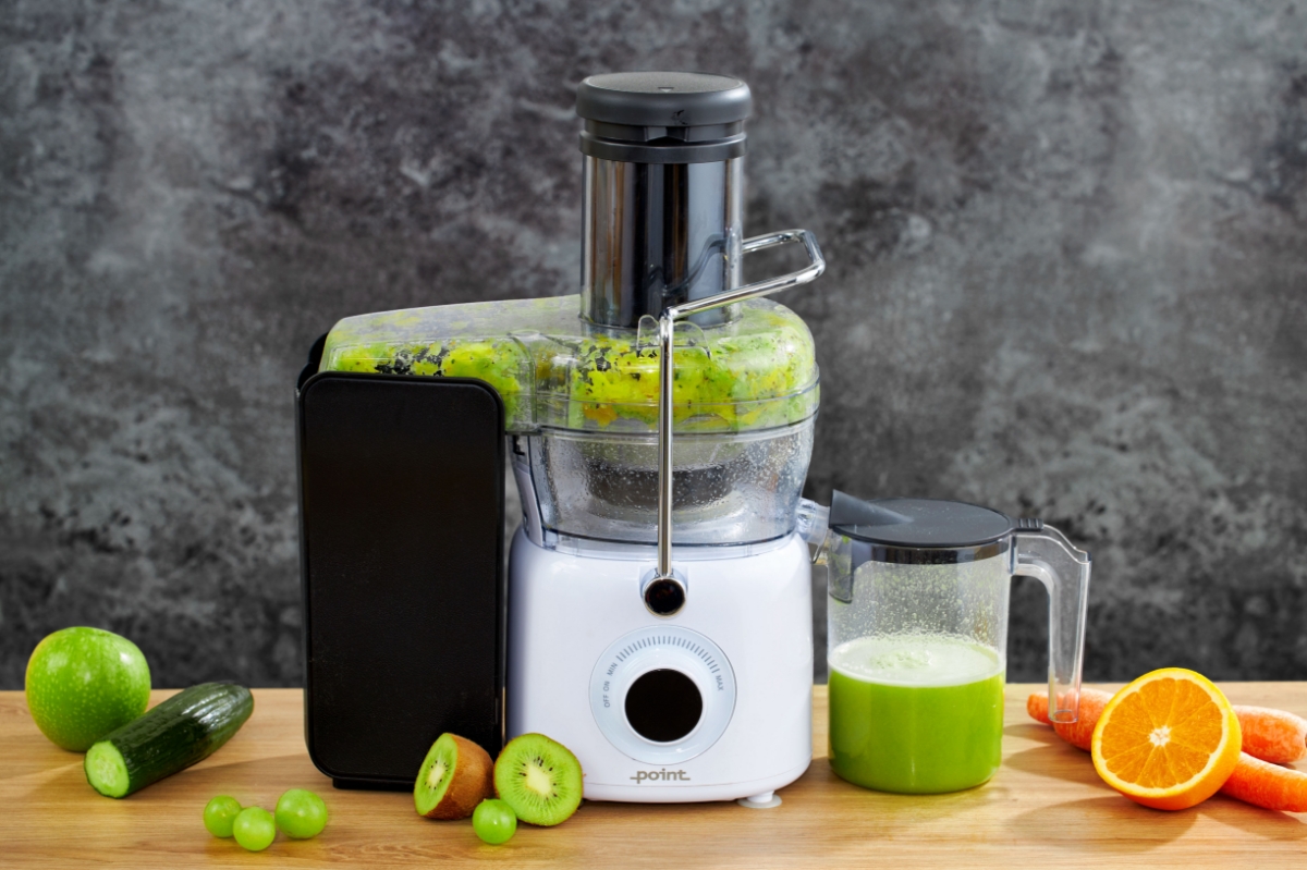 A white Point juicer on a wooden table with green and orange colored vegetables around it and a green juice inside the device