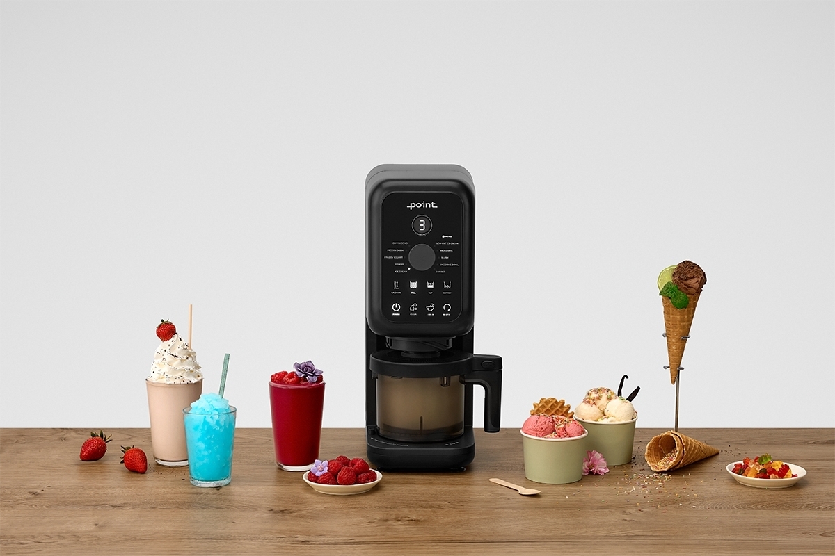 A black-colored Point ice cream machine on a wooden table with a light grey background with table full of different cold desserts next to the machine