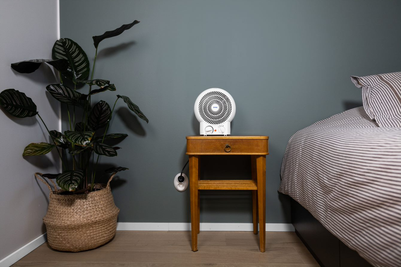 Senz SEFH20BA fan heater on a wooden bedside table, in front of a dark green wall, on the left is a tall plant in a woven basket. On the right is a bed with grey linen. 