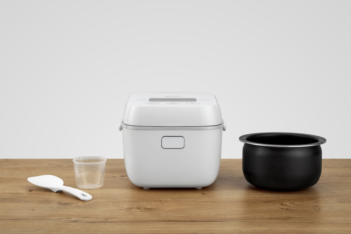 A white Point rice cooker on a wooden table with its inner black pot attached from the machine and placed next to it on the table and measuring cup and measuring spoon also next to the machine on the table