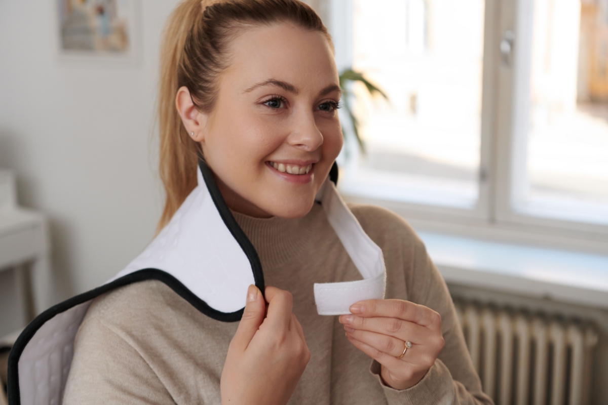 A smiling woman in a beige sweater placing a white Point heating pad to her shoulders