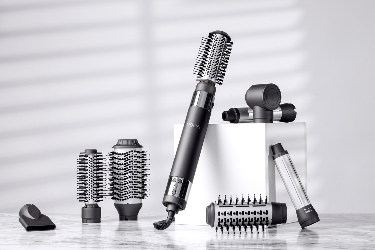 A black-colored NOOA Allure hot air styler placed on a white studio leaning against a white cube and with all its accessories next to it
