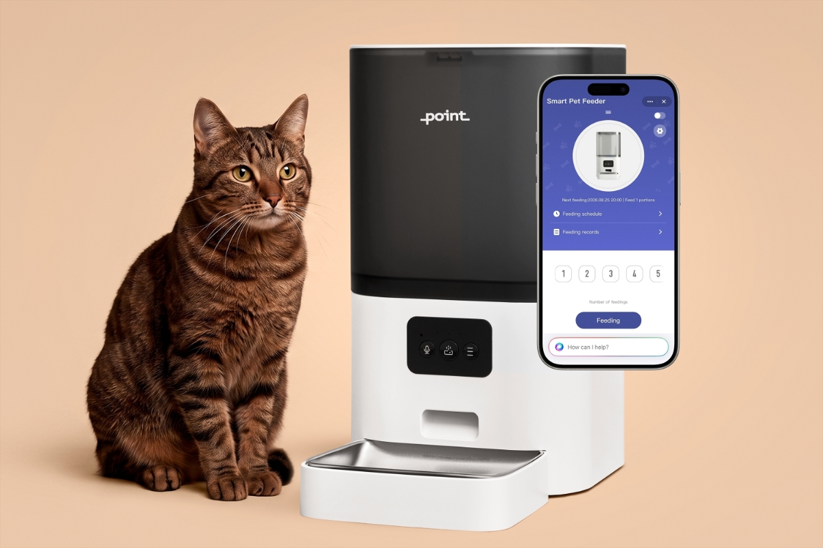 A brown cat sitting on a beige table looking near the camera and white Point Pet Feed smart feeder next to the cat and a smartphone in the air with an app opened on the screen