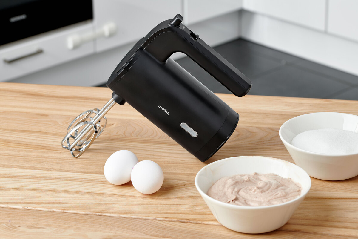 A picture of a black Point hand mixer standing on a wooden kitchen table with a bowl of dough and two eggs next to it on the table