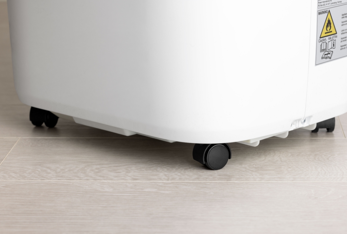 A close-up picture of the white Point air conditioner on a light wooden floor with its black wheels showing