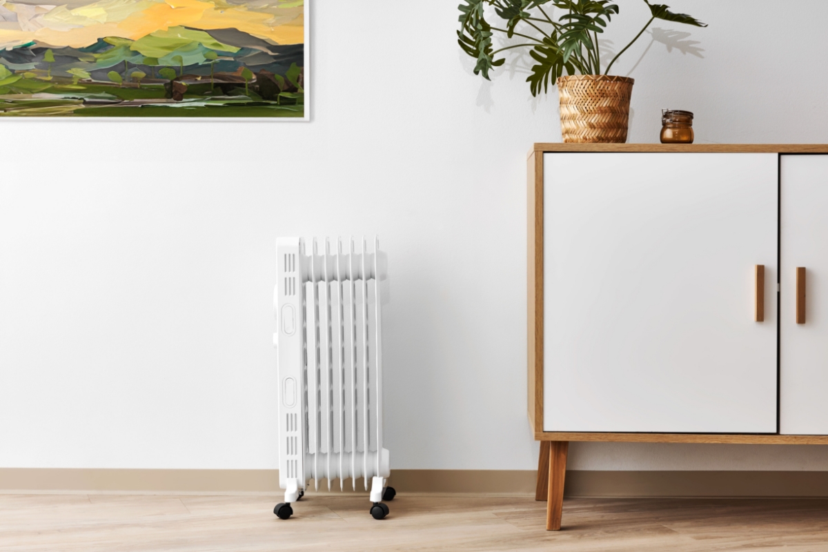 A picture of a white Point oil heater with black tires standing on a wooden floor and a wooden and white drawer next to it