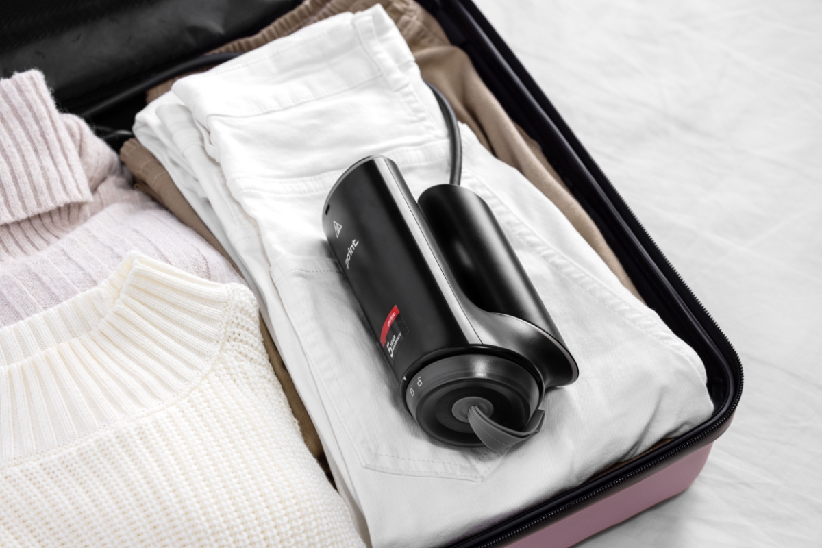 A black Point travel steamer folded and placed on a pink suitcase on top of white clothing ready to be packed on a trip