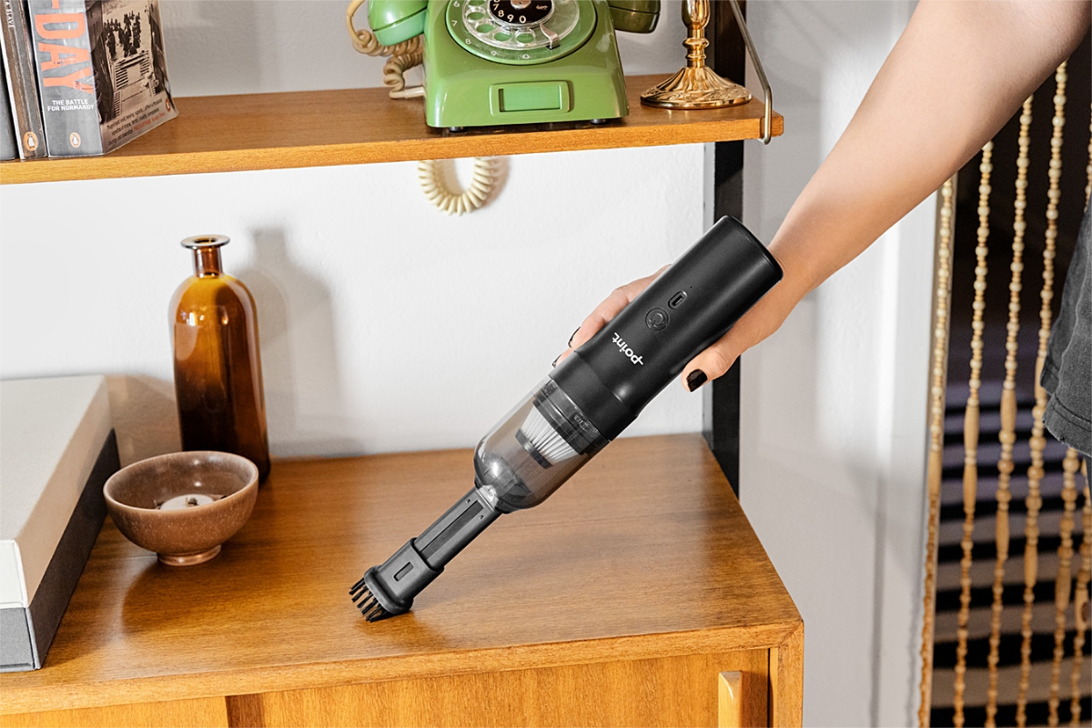 A black Point Tornado handheld vacuum cleaner on a person's hand vacuuming the top of a wooden drawer with retro glass bottles and an old telephone placed on the drawer
