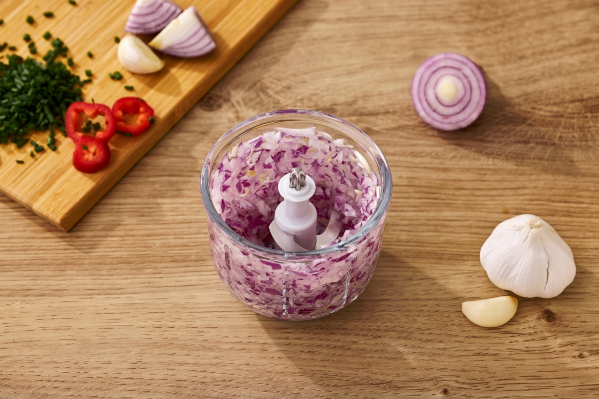 A picture of a Point cordless food chopper on a wooden table with its lid detached and the glass bown filled with chopped red onions and more onions surrounding the device on the table