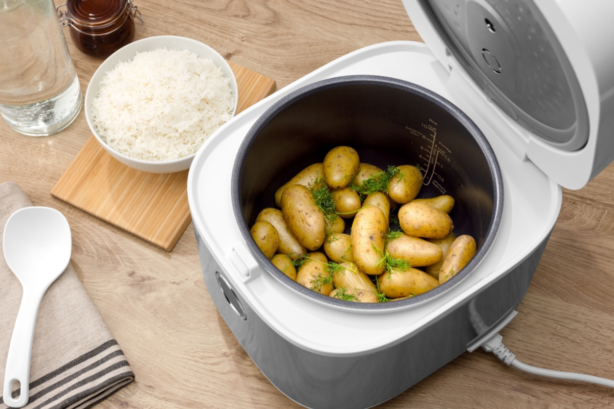 Close up of POINT PORC5010 RICE/MULTI COOKER 5L from above with the lid open. Inside are cooked potatoes with some dill. On the countertop is a bowl of cooked rice.