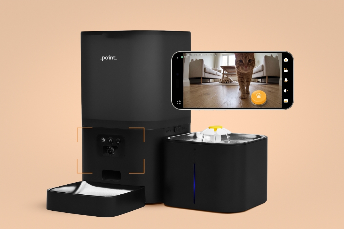 A black Point Pet Station smart feeder on a beige floor with a beige background and a smartphone in the air next to the device with a camera function on and a cat and a dog showing on the screen