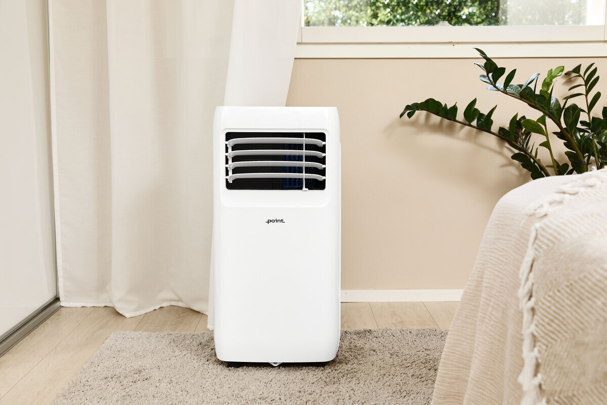 POINT POAC6022 air conditioner in front of a beige wall with a window on it, on the right corner is a green plant and a bed