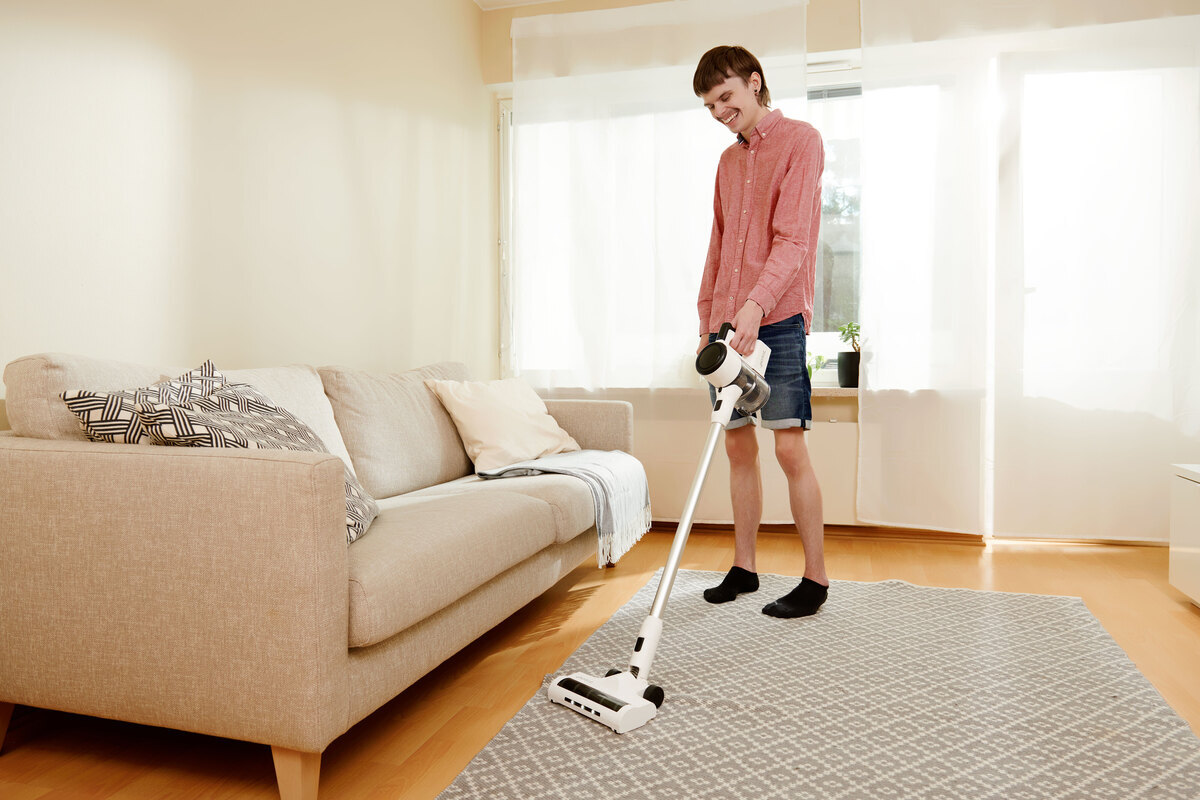 A wide angle image of a person in shorts and a light red shirt vacuuming a grey carpet with the Point Pencil stick vacuum cleaner