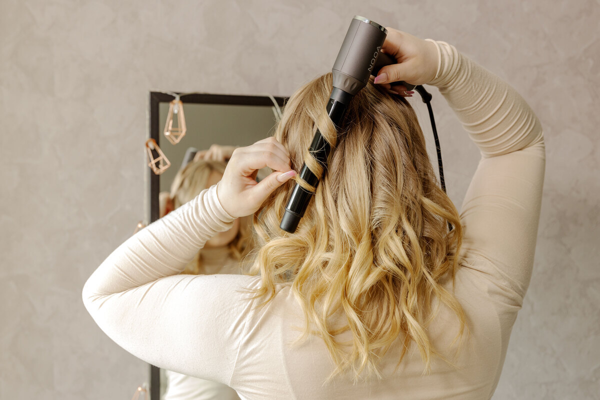 A person with blonde curled hair facing away from the camera, curling their hair with the Nooa Divine multi curler and a tapered curling wand on it