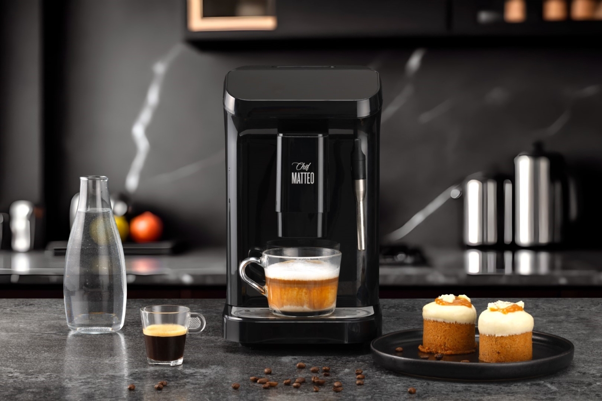 Wide angle image of CHEF MATTEO KEOPI COF.MACH BK/20BAR on a dark marble-like countertop, with two pastries next to it and a freshly brewed coffee under the spout