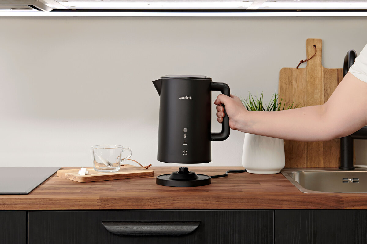 Wide angle image of the black POINT PRO POKED6015E KETTLE on a dark wooden kitchen counter as a person lifts it off the base