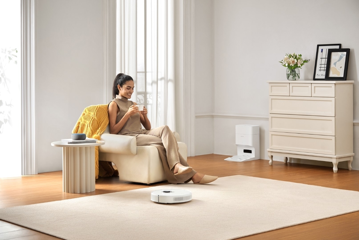 DREAME D10 PLUS GEN2 ROBOT VACUUM WH vacuuming a beige carpet as a person sits in an armchair next to it drinking from a mug