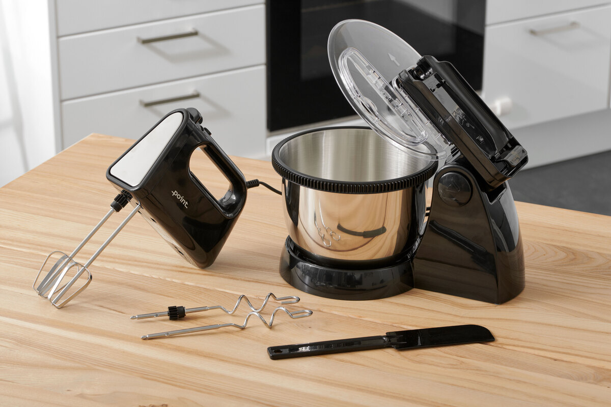 A picture of a black and steel hand mixer with a bowl standing on a wooden kitchen table with all its accessories next to it on the table