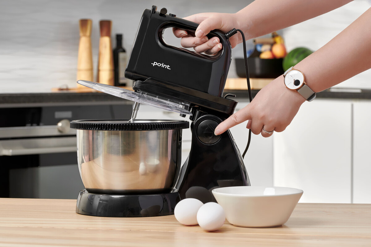 A picture of a black and steel hand mixer with a bowl standing on a wooden kitchen table with a person adjusting its whisk and two eggs on the table next to the appliance