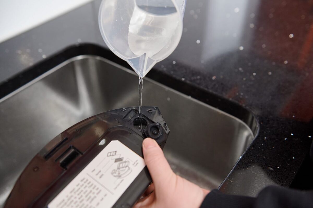 A picture of a black Point Dusty robot vacuum cleaner's water tank being filled with water with a plastic cup on top of a sink