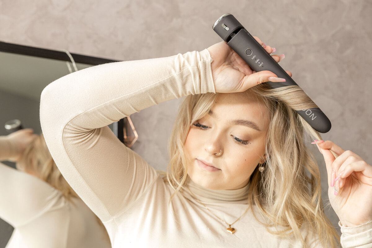 A woman straightening her blonde hair with the dark grey NOOA straightener and a mirror behind her