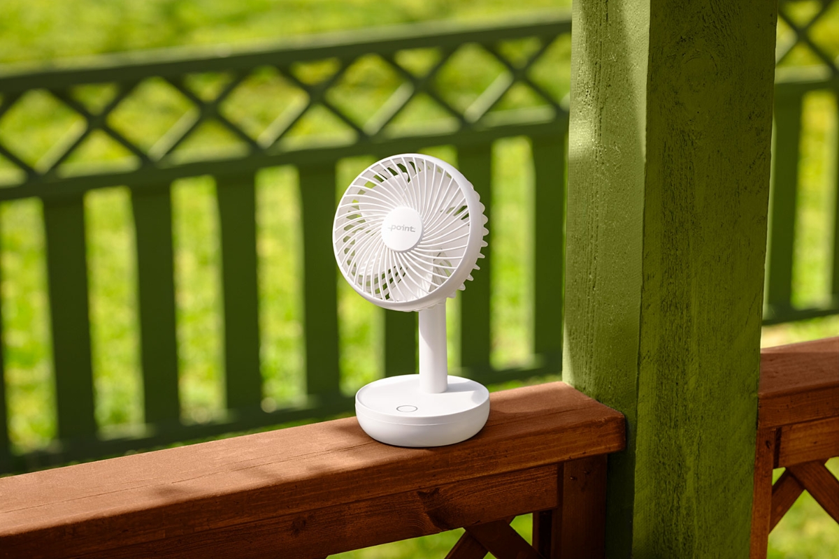 A white and small Point portable table fan on the edge of a wooden balcony with lots of sun and a green yard showing in the background