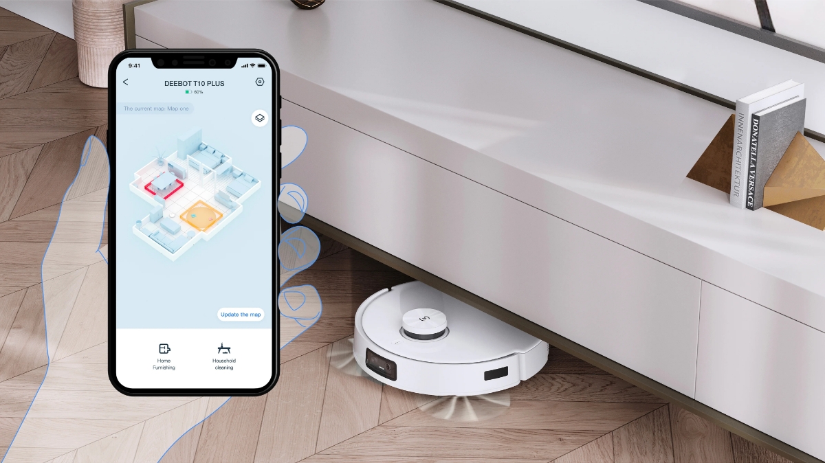 An Ecovacs robotic vacuum cleaner cleaning under a shelf and a hand holding a smartphone with the Ecovacs app next to it