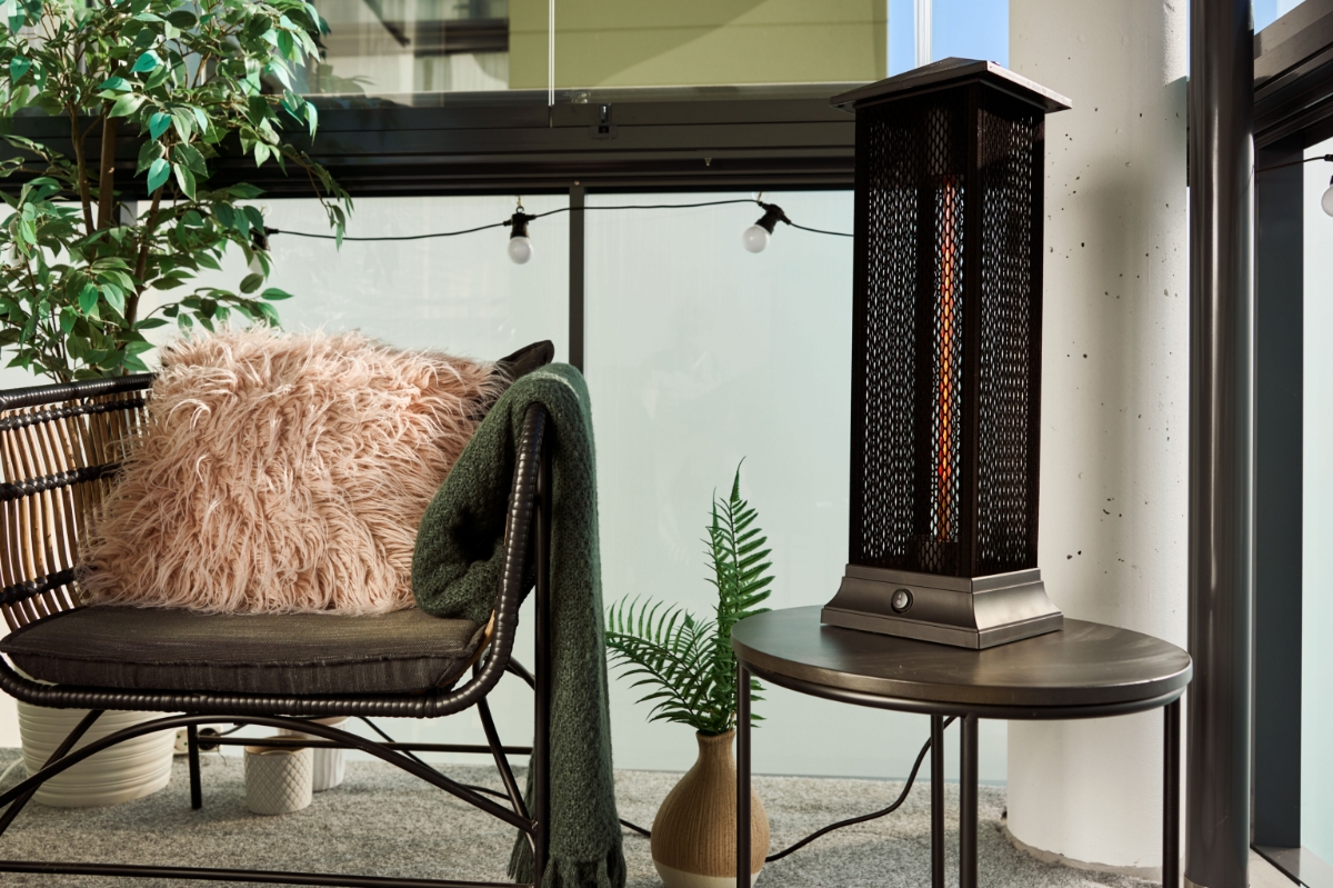 A wide image of the POINT PRO POPHTOW66 patio heater on a side table, on a sunny balcony. On its left there is a lounge chair with a cozy pillow and throw blanket as well as some plants