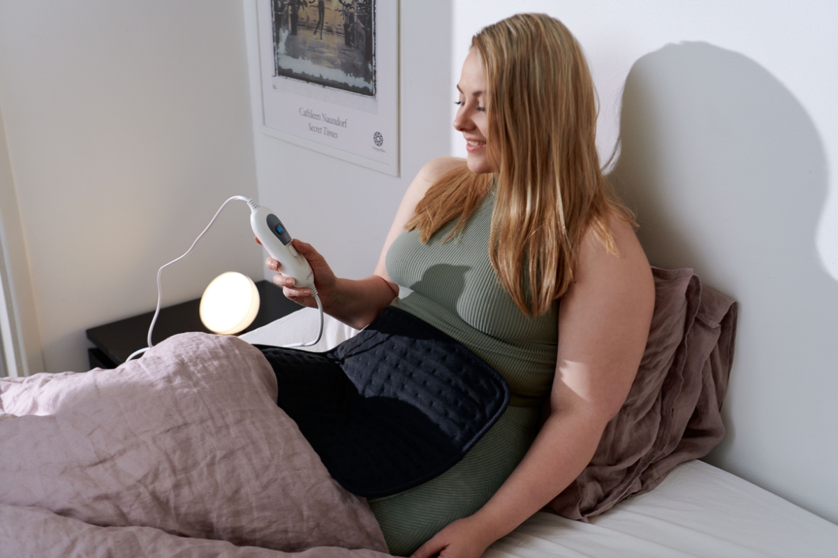 A woman in a green shirt holding a black Point heating pad on her stomach while laying on a bed with pink sheets and holding the remote control in her other hand