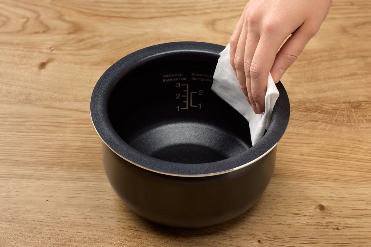 The black inner container of the Point rice and multicooker placed on a wooden table and a hand wiping it clean with a white cloth