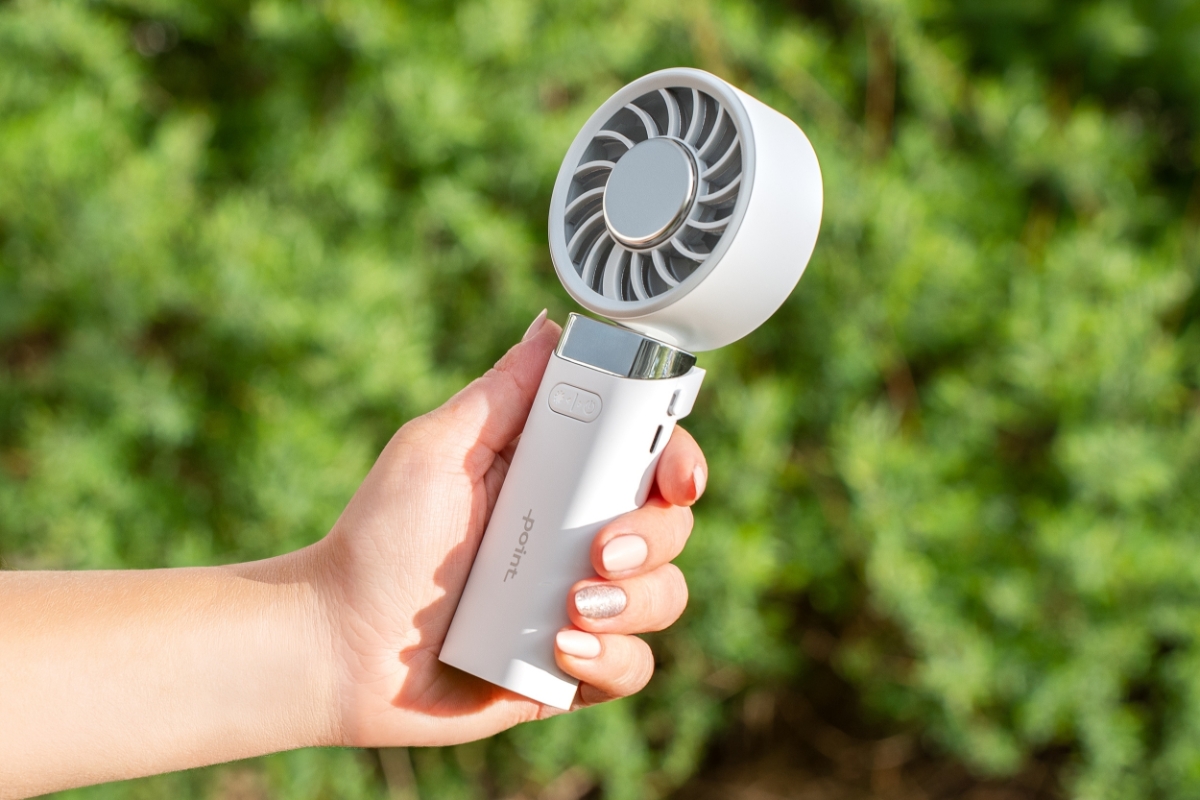 POINT HANDHELD FAN WH A white Point hand fan being held in a person's hand and a green bush behind the fan