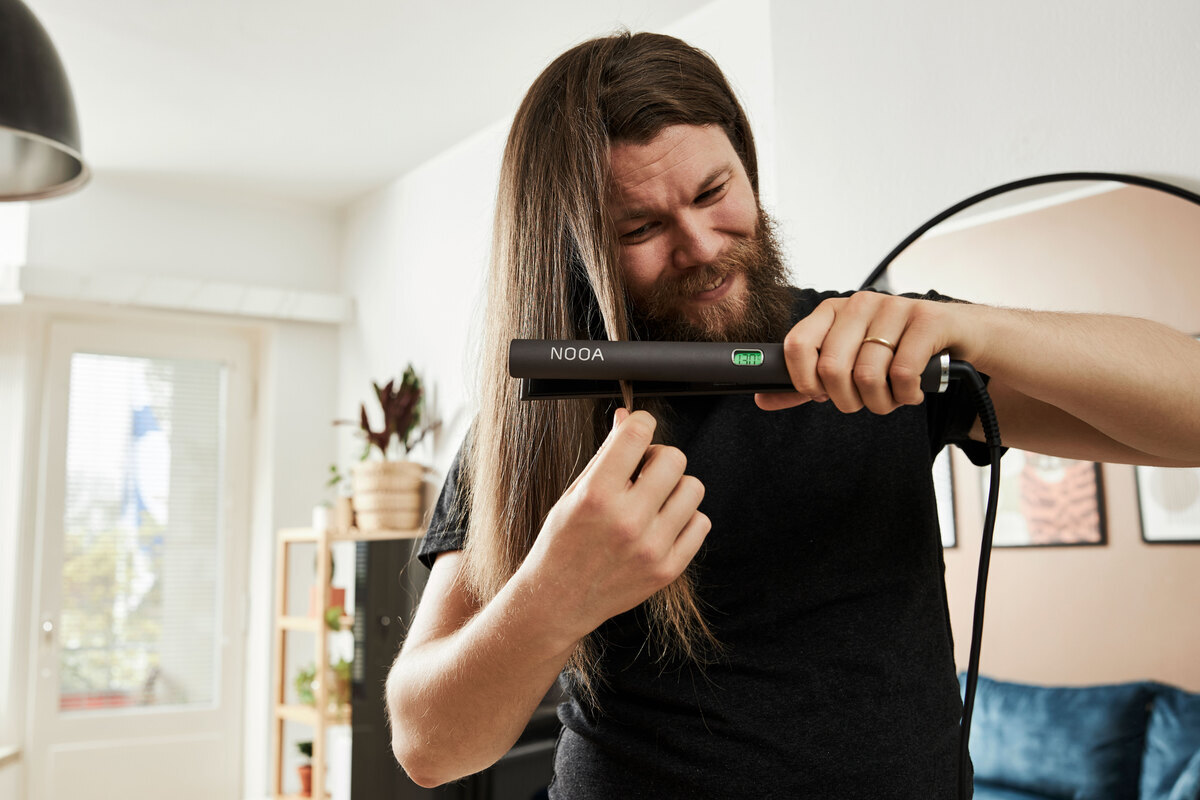 A man straightening his brown hair in a living room with a grey NOOA hair straightener with a mirror behind him