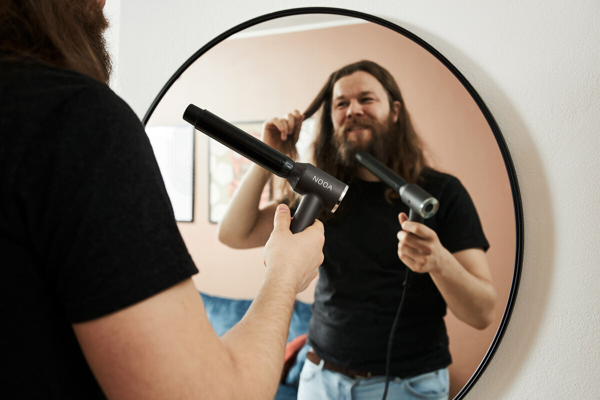 A person with thick brown hair wearing a black t-shirt is looking at themselves in the mirror, about to use the Nooa Divine multi curler on their hair