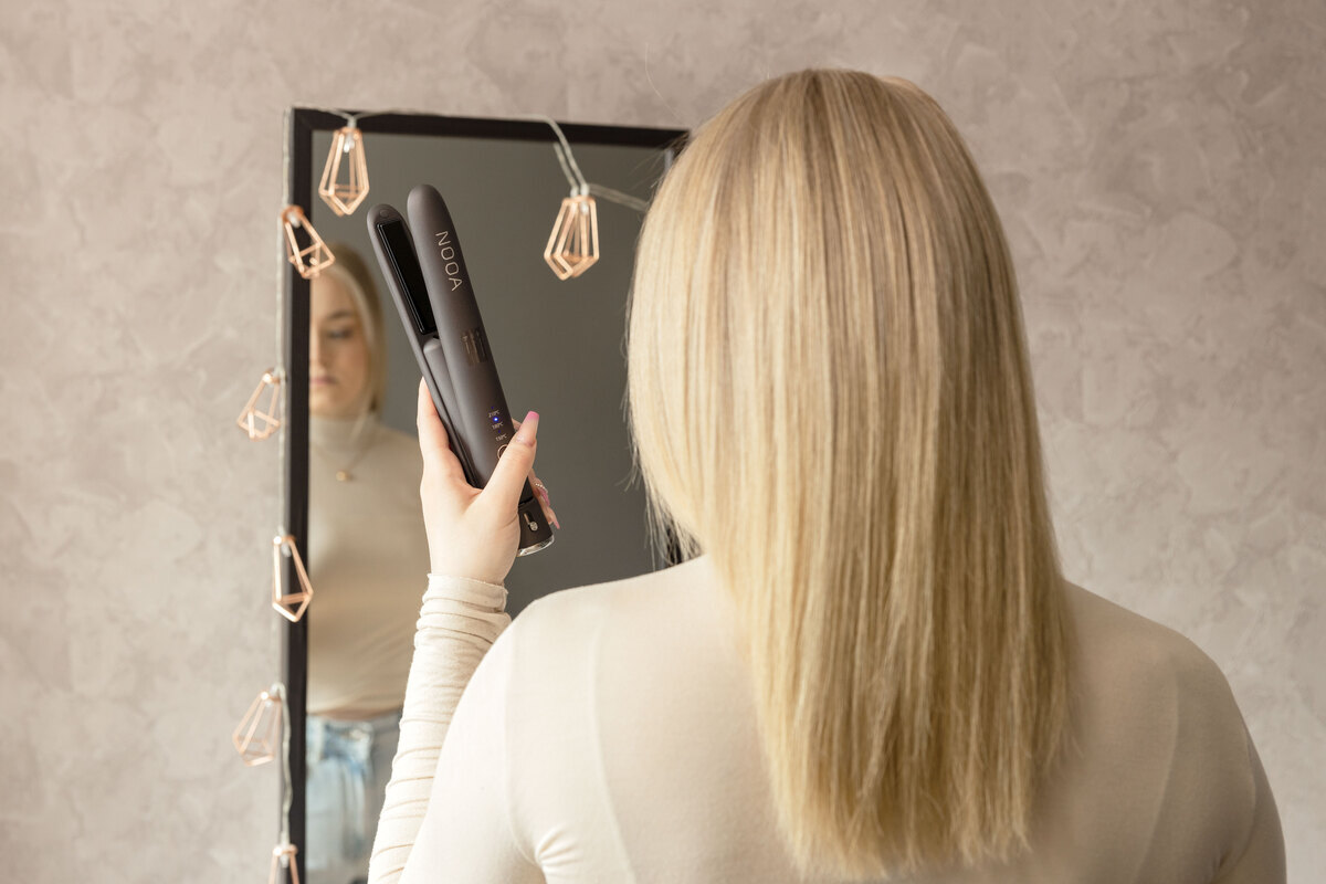A woman holding a dark grey NOOA straightener in front of a mirror and a pink wall