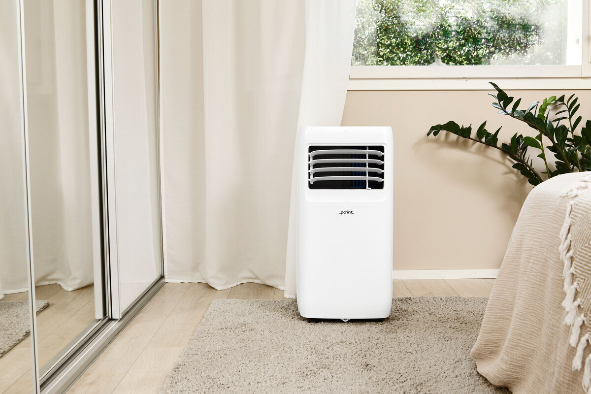A wide angle picture of the POINT POAC6022 air conditioner in front of a beige wall with a green plant behind it next to the wall 
