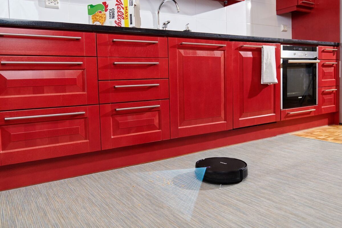 A picture of a black Point Dusty robot vacuum cleaner vacuuming the floor of a kitchen with red cabinets from spilled cereal and the infraded sensor showing