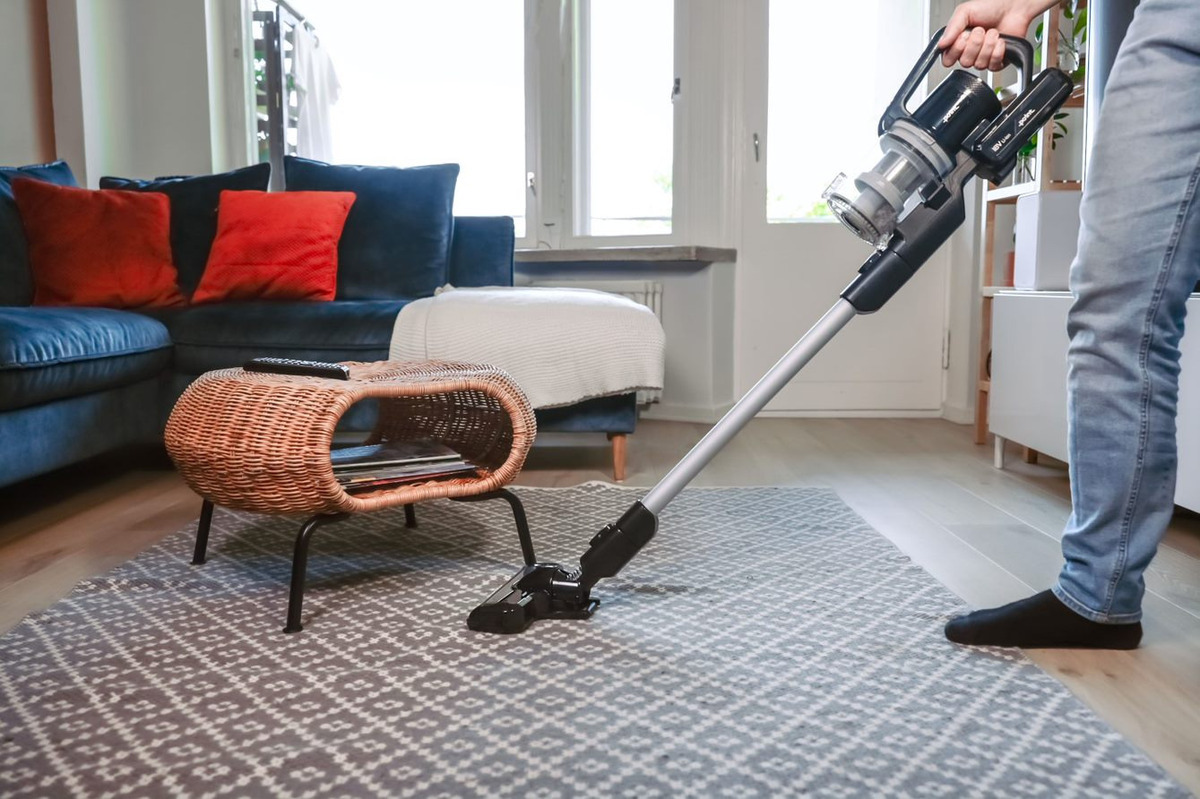 A person with blue jeans vacuuming underneath a woven coffee table with the POINT POVC618DB 18V stick vacuum in a living room with a blue sofa and grey patterned carpet