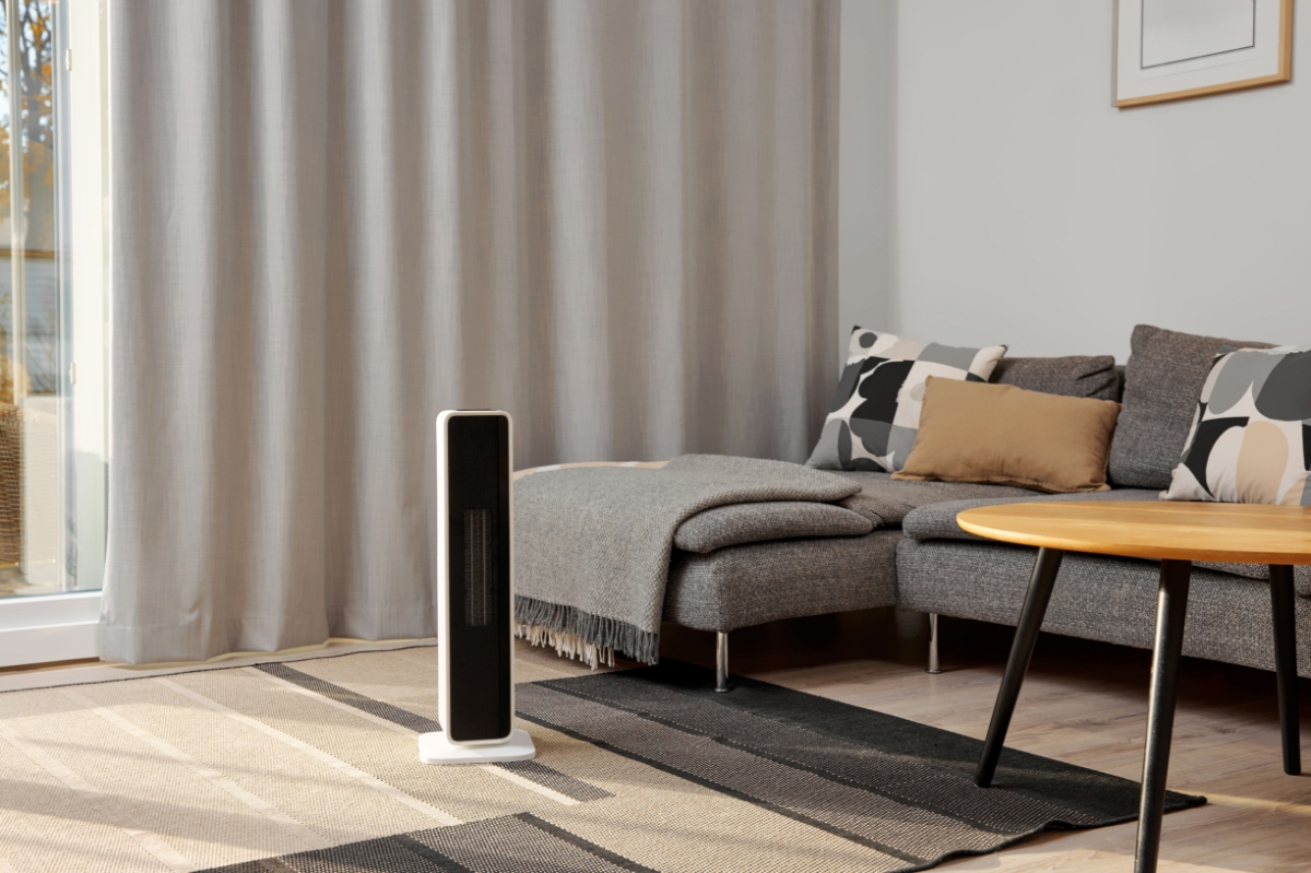 A black Point fan heater standing in a living room with beige and black carpet and a grey sofa next to the heater and light-grey curtains behind the appliance
