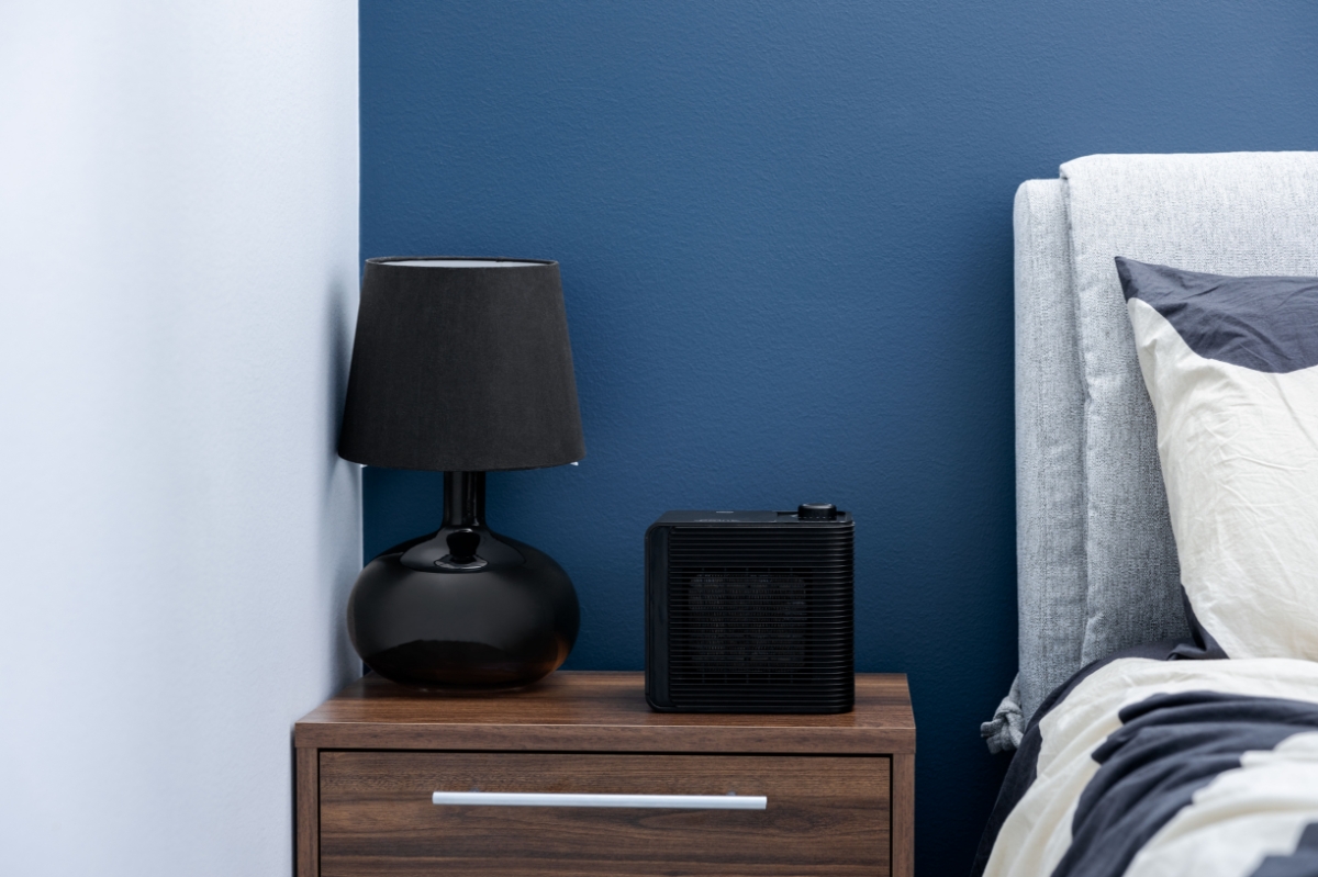 A black Point fan heater standing on a dark wooden table next to a black lamp and a bed with grey-white sheets