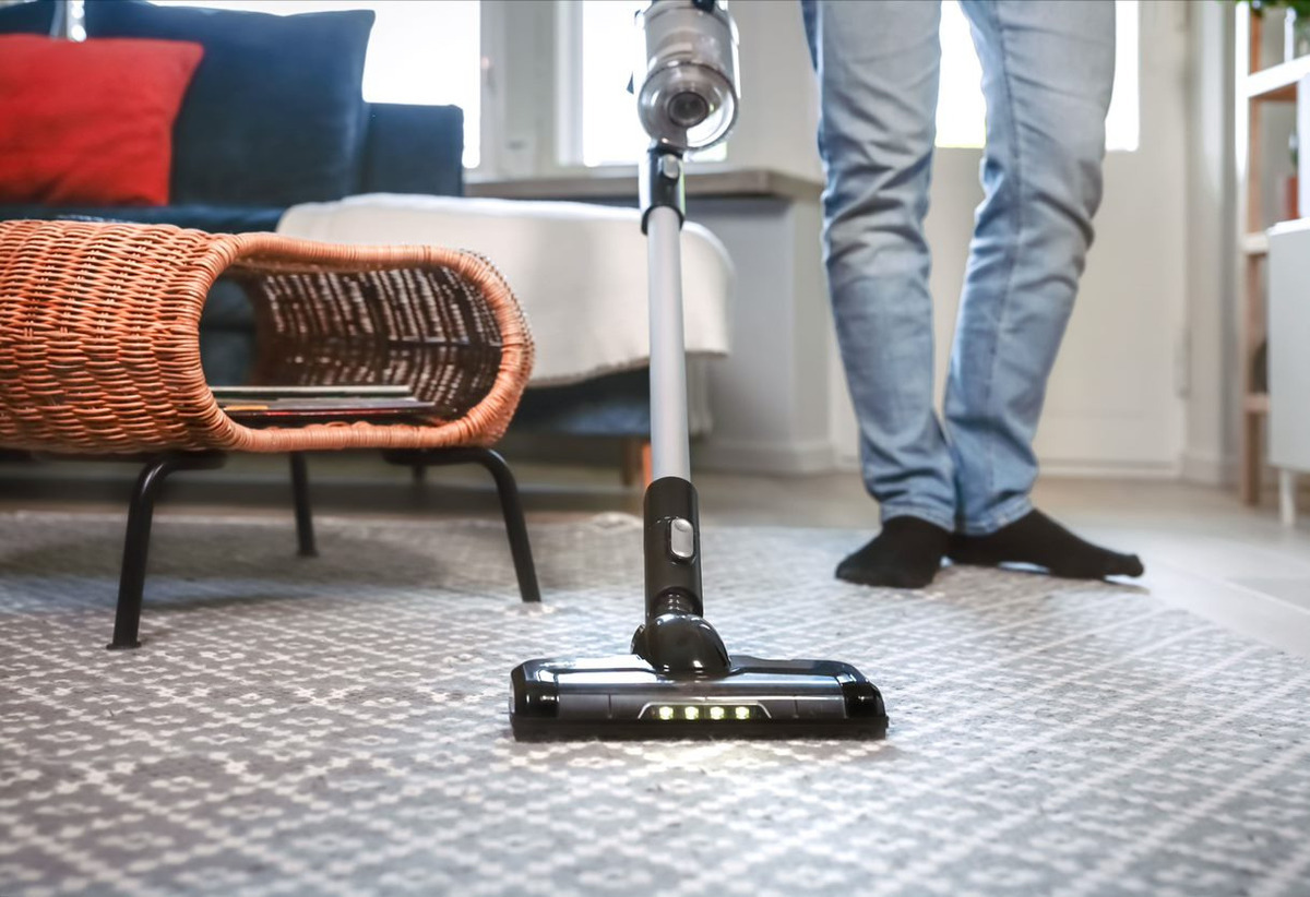 Close up of the POINT POVC618DB 18V stick vacuum on a grey patterned carpet, in front of a blue sofa and brown coffee table, a person with blue jeans holding it