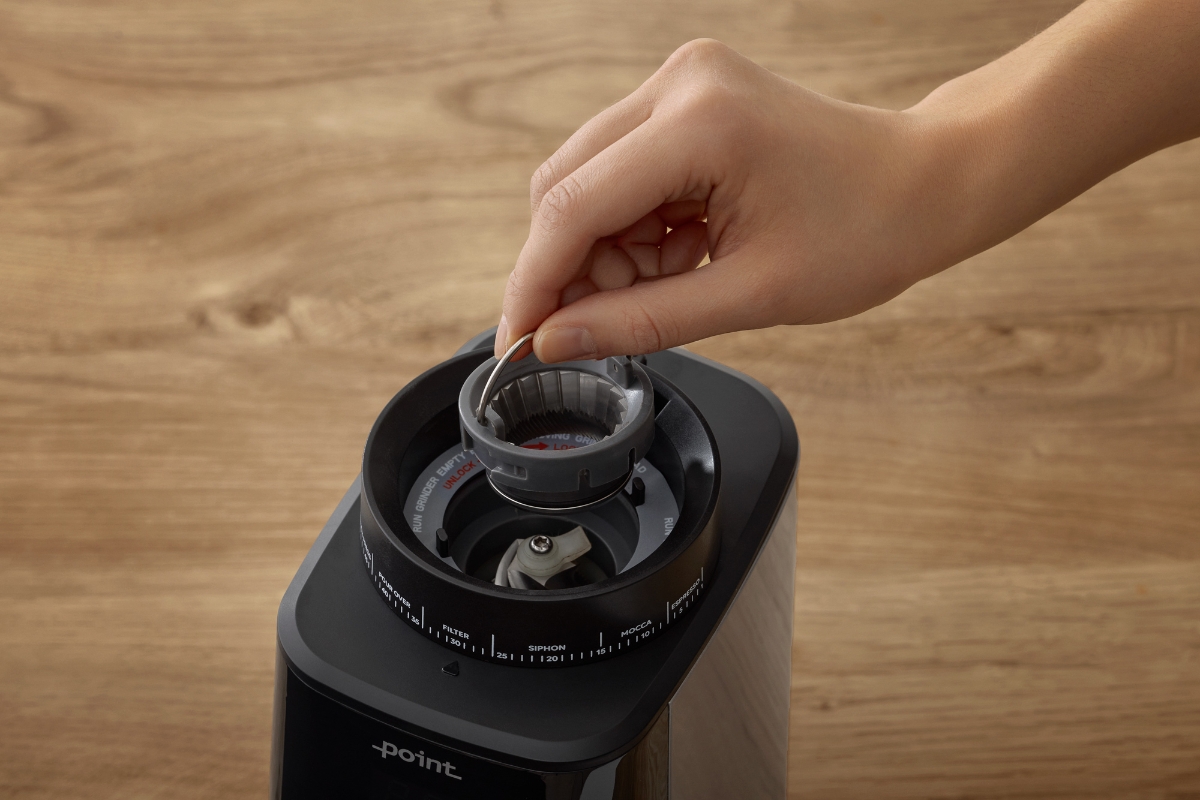 A black Point Pro coffee grinder pictured up close with a person holding its grinding adjustment roller placed inside the machine and a wooden table showing in the background