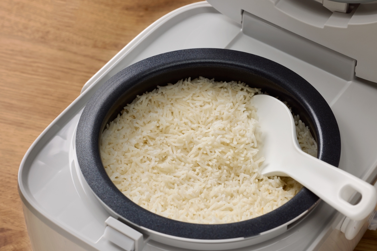 A white Point rice and multicooker filled with ready made white basmati rice and a measuring spoon among the rice and the device on top of a wooden table