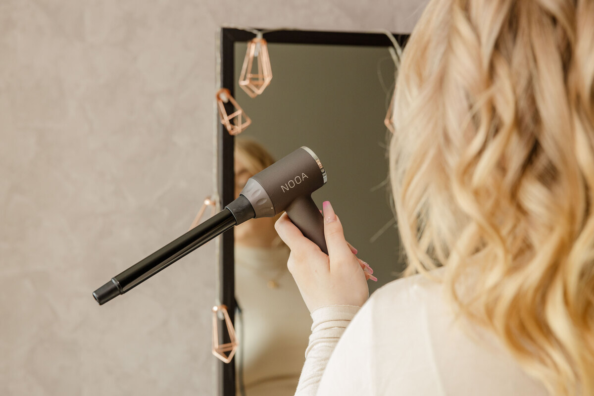 A person with blonde curly hair is facing away from the camera, towards a mirror, holding the Nooa Divine multi curler in their hand