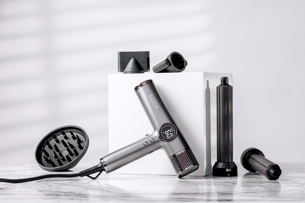 A studio picture of a matte grey NOOA hair dryer next to a white cube and all its accessories surrounding the hair dryer