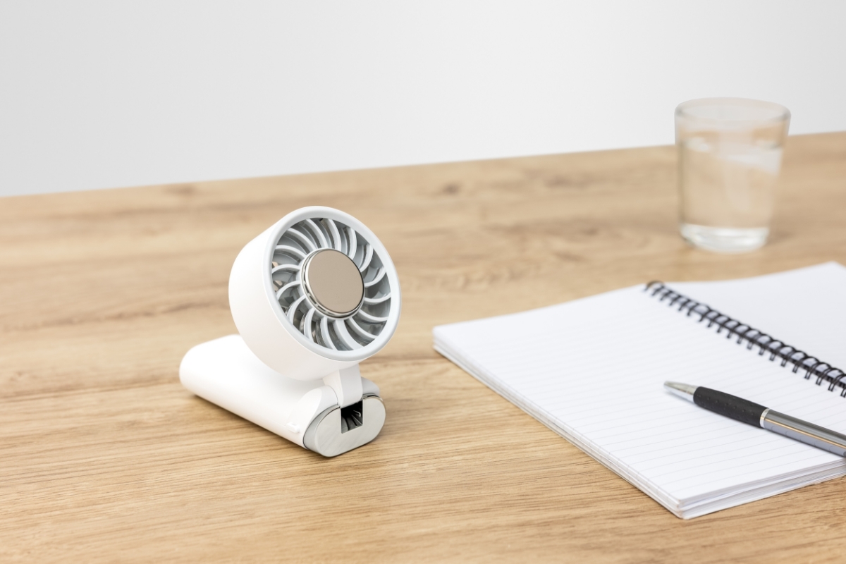 POINT HANDHELD FAN WH A white Point hand fan on a wooden table in folded position pushing air towards the air and a writing paper next to the fan on the table